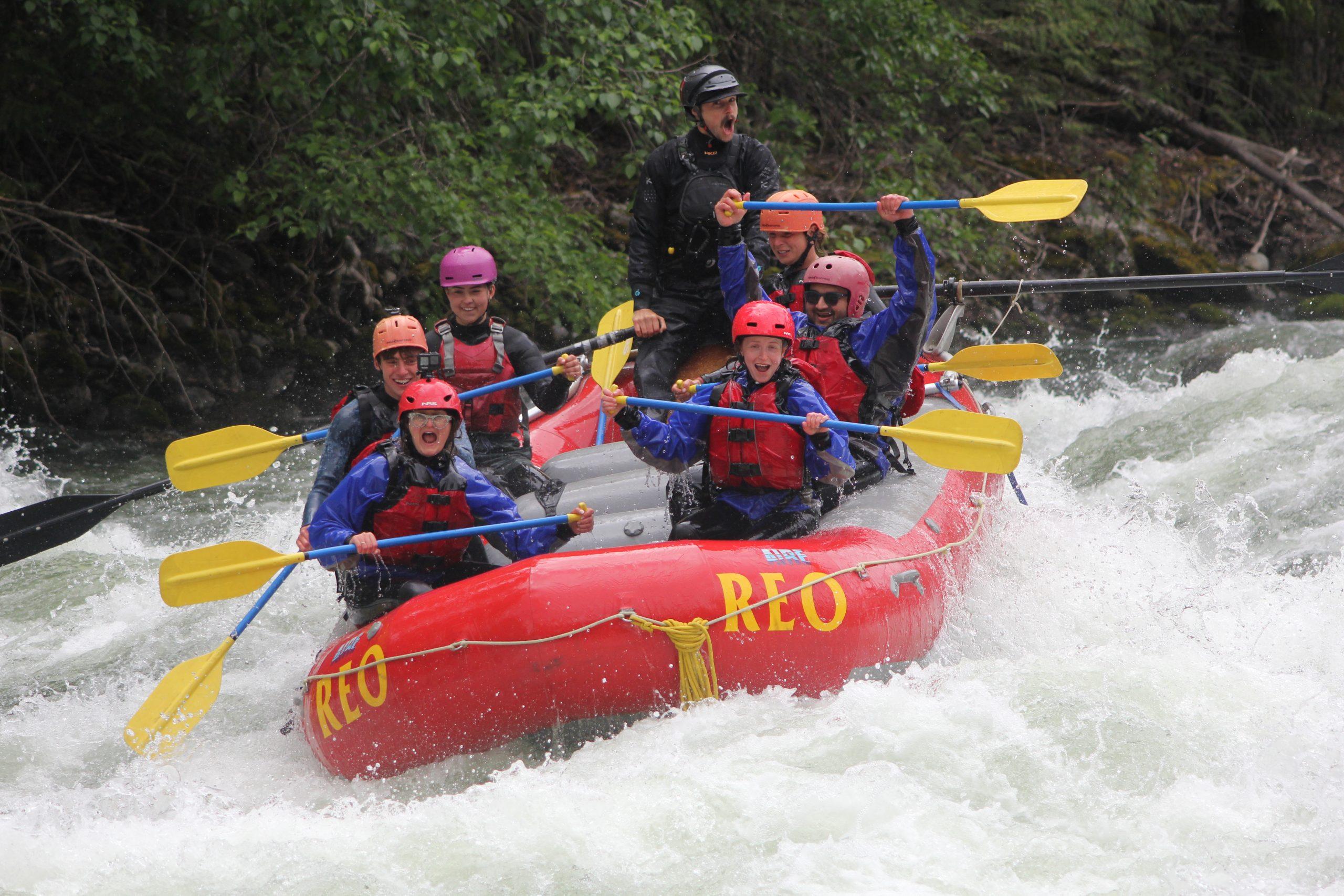 A group of seven rafters paddle through whitewater rapids on the Nahatlatch River in a red REO Rafting raft, grinning and laughing as water splashes around them.